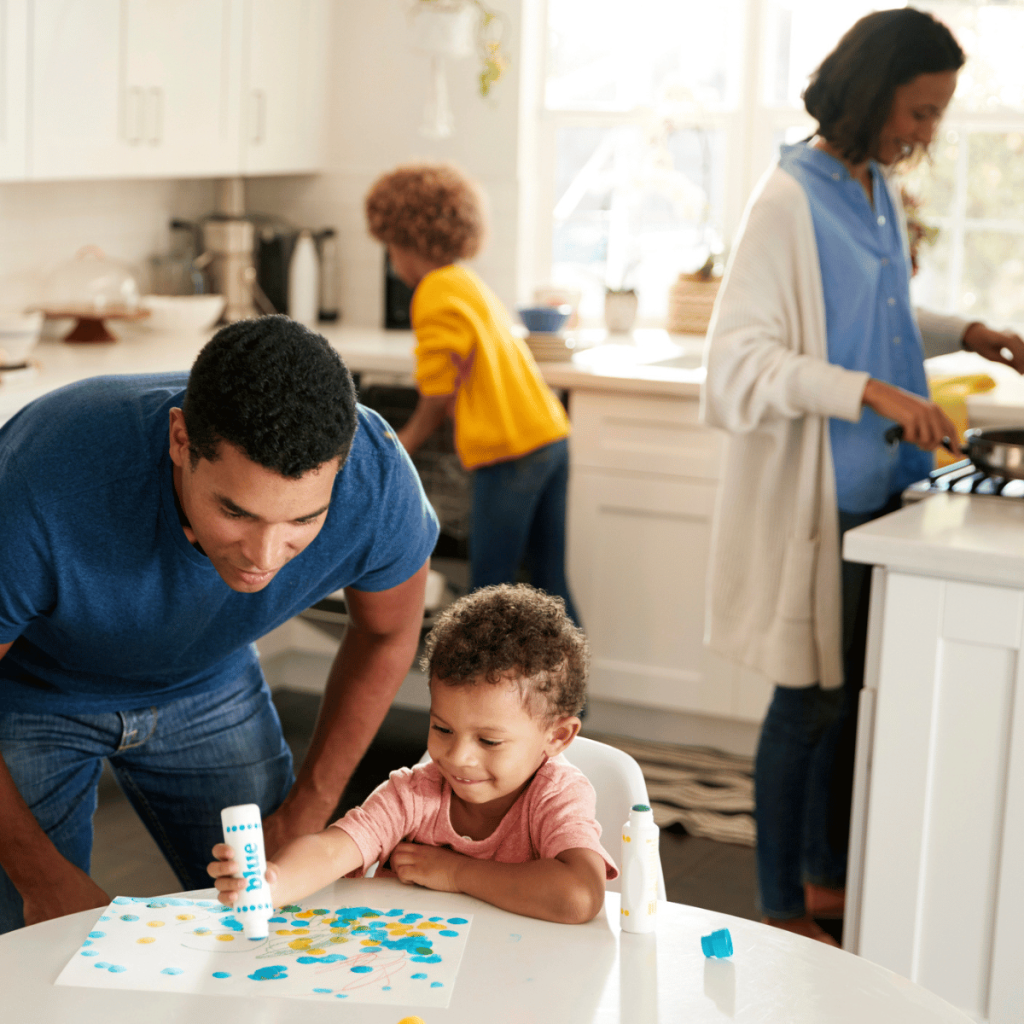 family in kitchen: one child is helping with dishes, mom is cooking, dad and the other child are doing a learning activity at the table