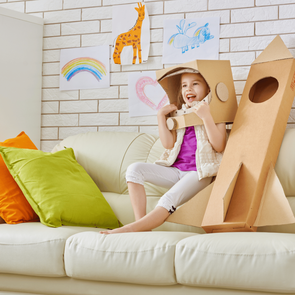 young girl on couch with a spaceship and helmet made out of cardboard boxes